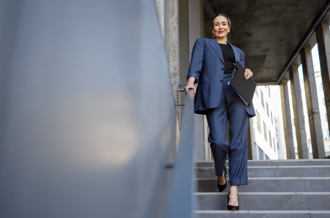 Pretty business woman with clipboard folder walks down the stairs near the office building and looks at the camera